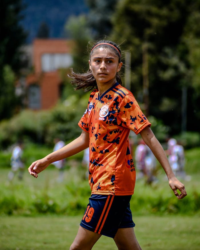 A young female athlete in an orange jersey plays on an outdoor football field.